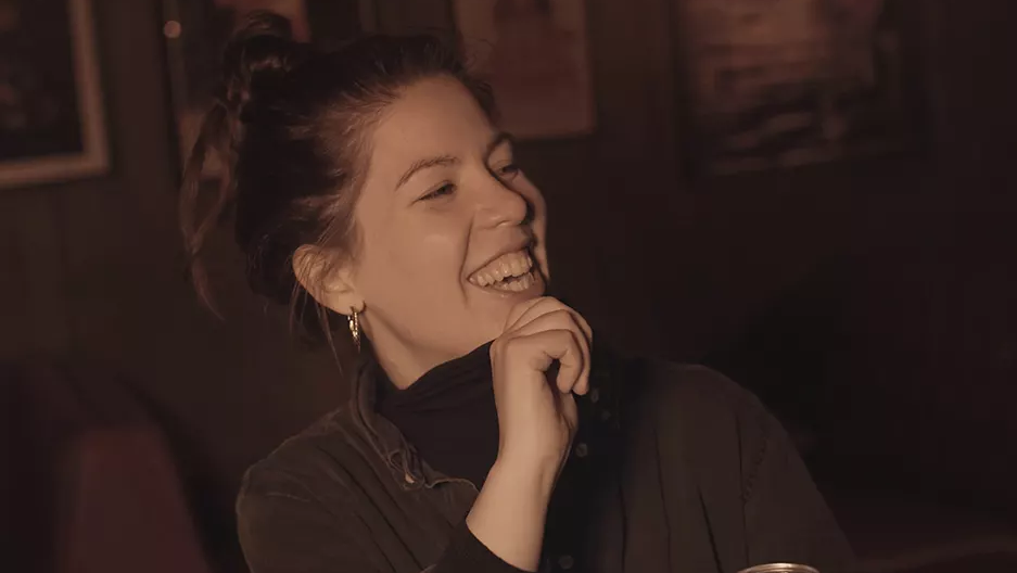 woman smiling and laughing in a dark bar, holding the collar of her jumper