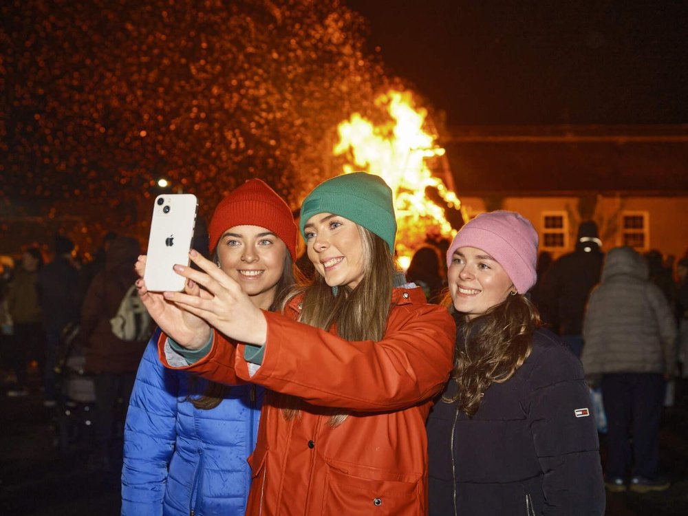 Three young women take a selfie at a nighttime festival with a fire sculpture in the background.