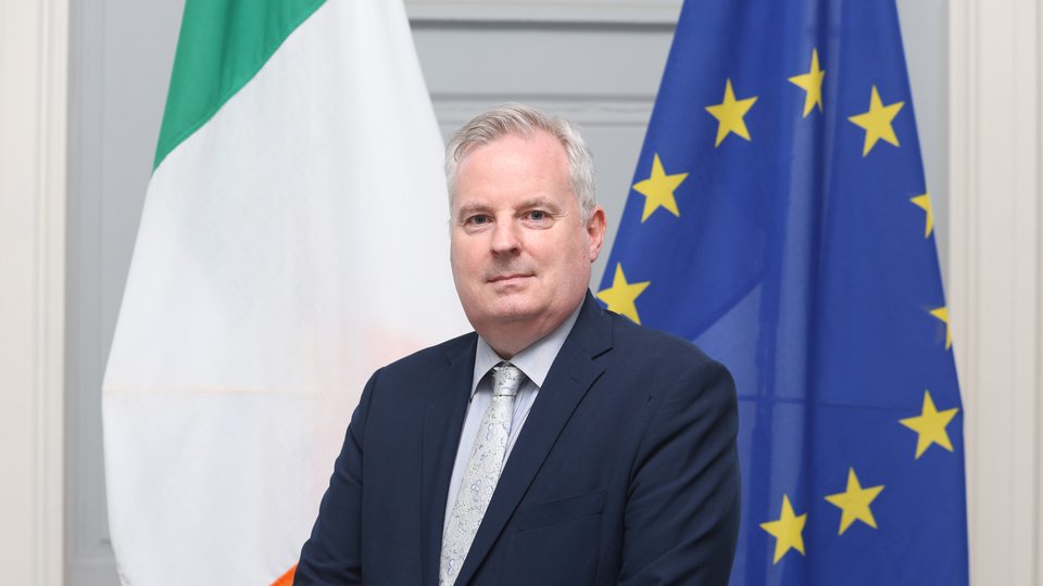 Consul General Gerry Kelly headshot in front of Irish and EU flags