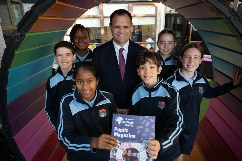 Man standing in a semi circle with six school children gathered around him and holding a magazine