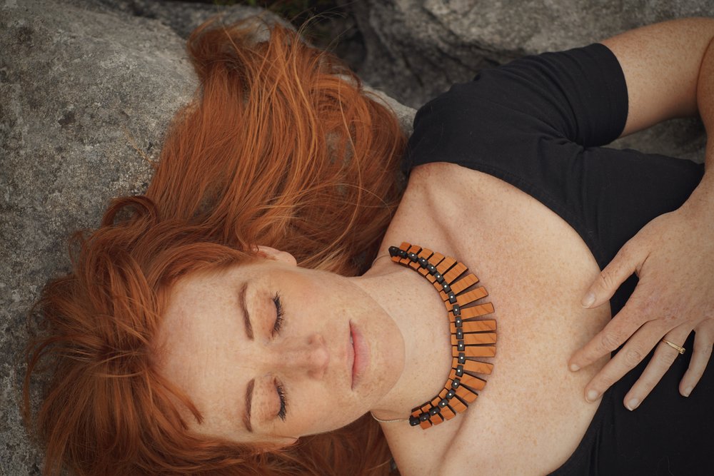 A woman lies with her eyes closed on craggy stones. The warm tones of her wooden necklace, creating a torc shape with different lengths of rectangular wood are accentuated by her bright red hair