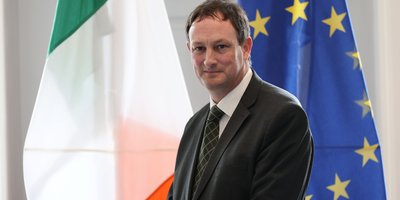 A man in a suit standing in front of the Irish and European flag