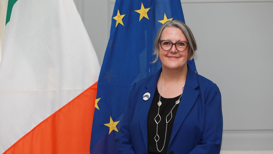 Headshot of Michelle Winthrop standing in front of the Irish and EU flags.