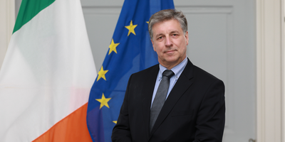 Ambassador headshot in front of Irish and EU flags.