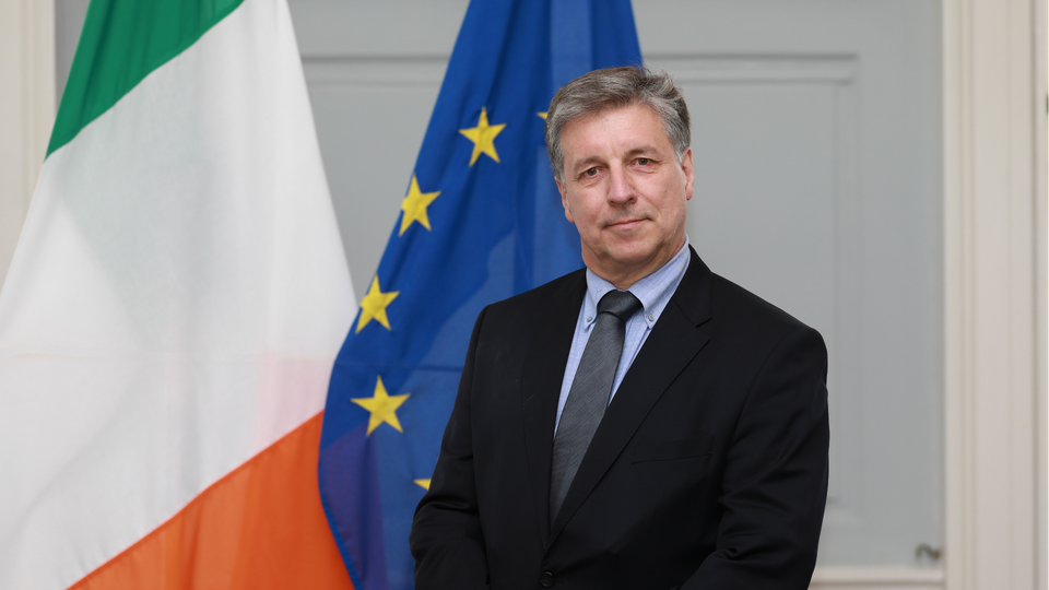 Ambassador Patrick Empey standing in front of the Irish and EU flags.