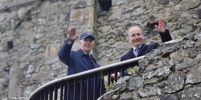 two men on a balcony waving, with a stone wall behind and in front of them
