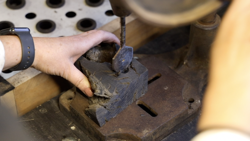 Close-up image shows a woman using a drill press, her left hand is holding bog oak on the machine's metal base.