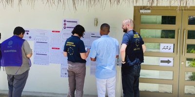 Election observers in EU-branded polo shirts stand outside a polling centre with pages covering the wall showing tables and other text.