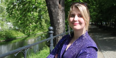 A headshot of a blonde woman, wearing purple with her arms crossed, smiling.
