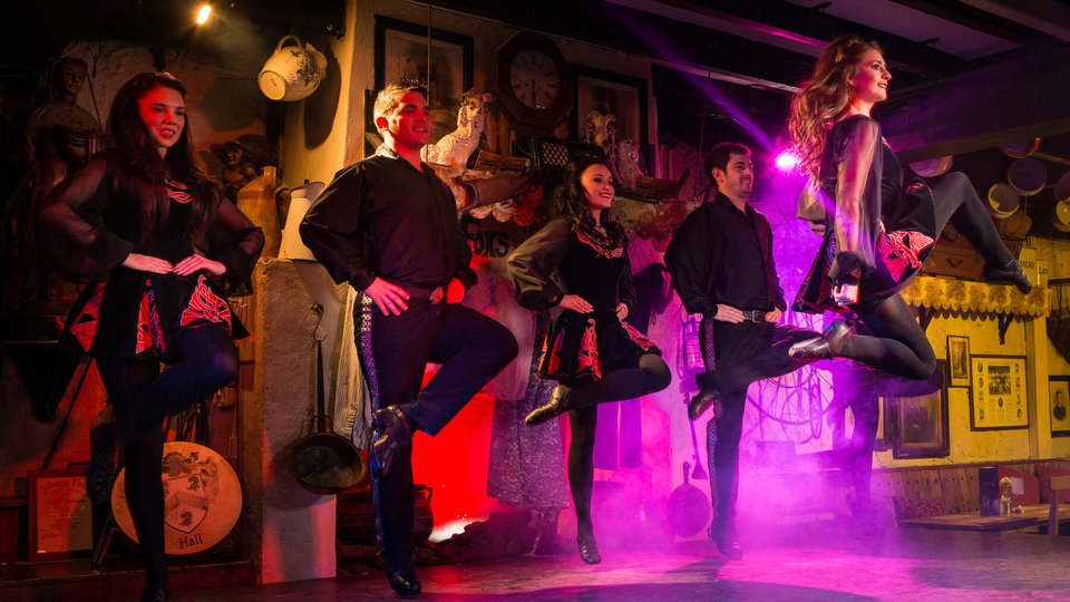 four irish dancers in the air with legs crossed in a dance with purple lighting behind them