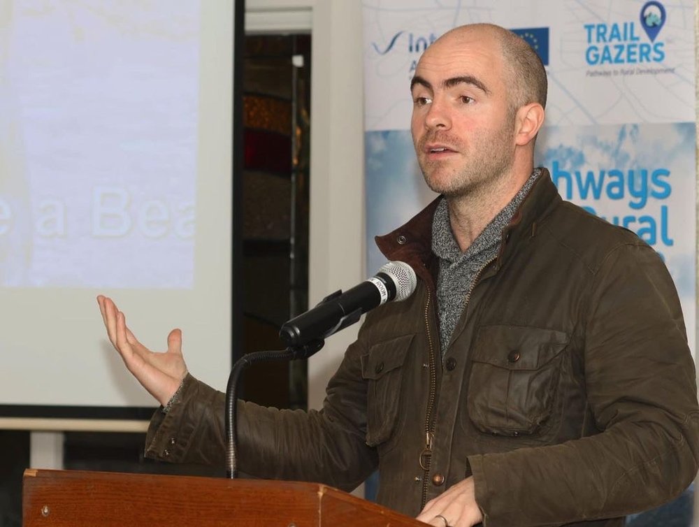 Killian McLaughlin gestures while speaking into a microphone at a podium. He is wearing a brown jacket and grey jumper.