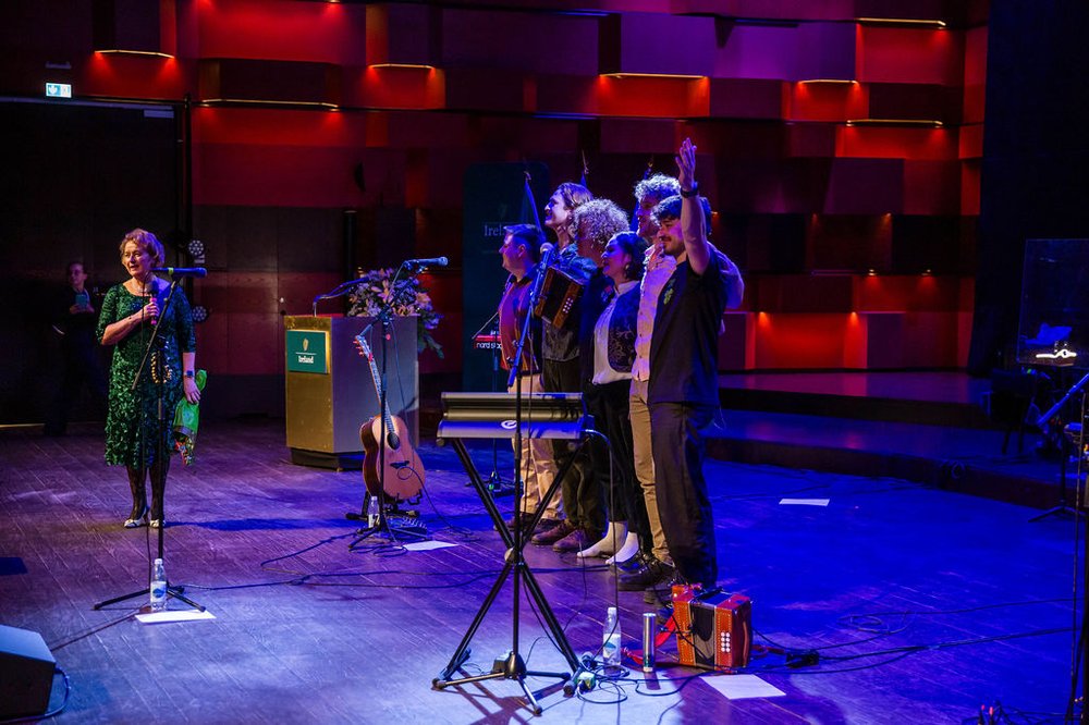 Ambassador Barbara Jones stands onstage with trad fusion band Moxie with their arms raised to the audience at the end of their performance.
