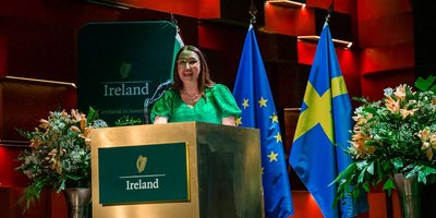 Minister Emer Higgins stands at the podium at the Royal College of Music in Stockholm at the Saint Patrick's Day concert. Behind the Minister are the flags of Sweden and the EU.