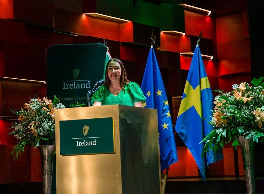 Minister Emer Higgins stands at the podium at the Royal College of Music in Stockholm at the Saint Patrick's Day concert. Behind the Minister are the flags of Sweden and the EU.