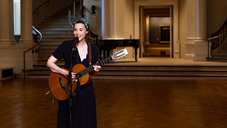 a woman playing guitar standing up in a grand hall with steps behind her