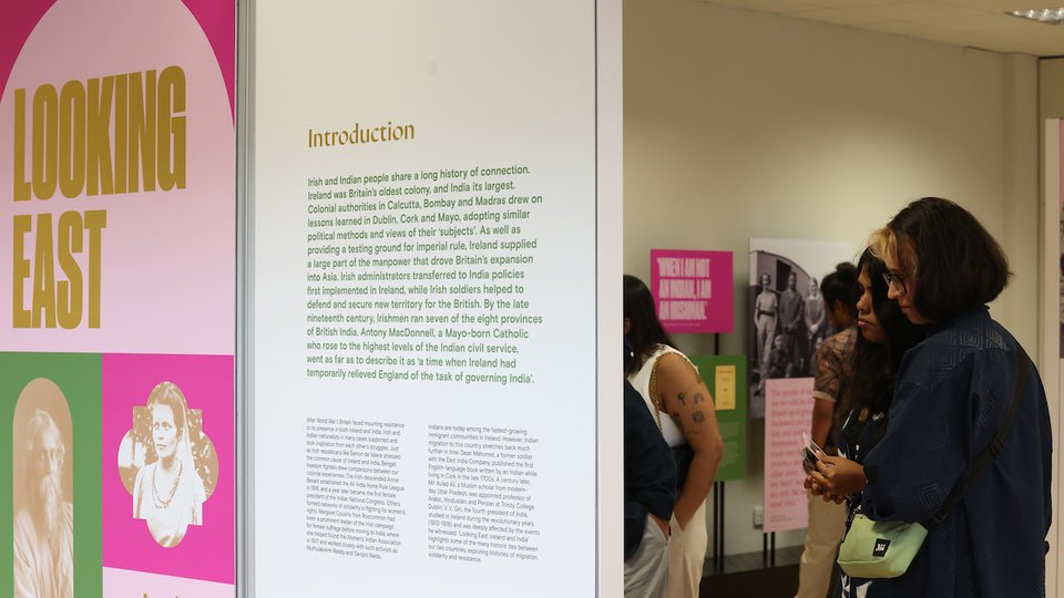 An exhibition panel with a pink, green and gold graphic reading: Looking East: Ireland and India, as a woman looks at the panel behind