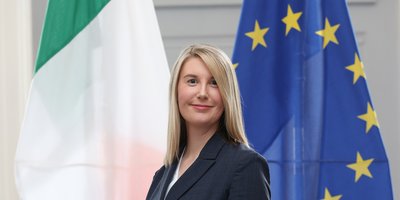 Maria Sheehy looking at camera in business suit, flanked by flags of Ireland and the EU
