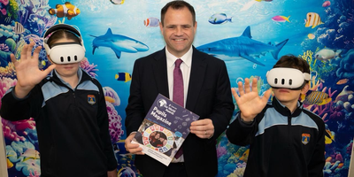Man in suit holding a magazine with two school children with VR sets on their heads and backdrop of ocean life behind them