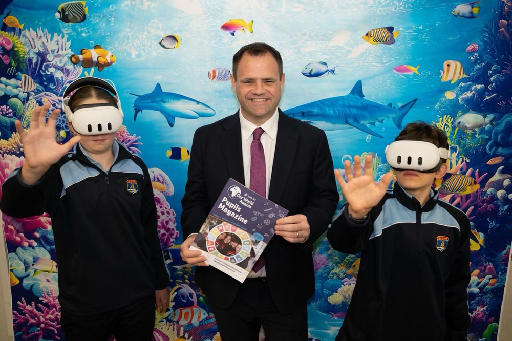 Man in suit holding a magazine with two school children with VR sets on their heads and backdrop of ocean life behind them