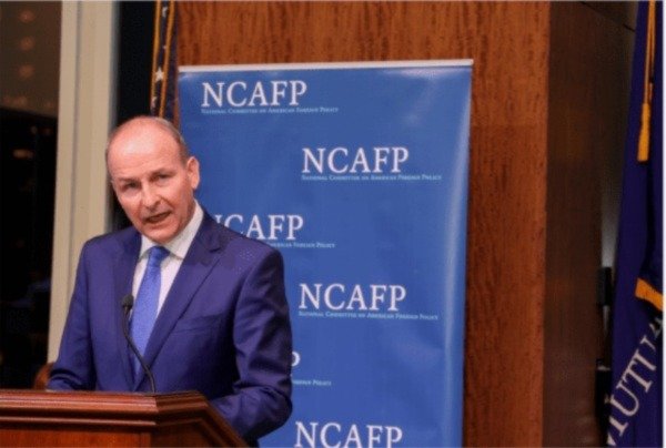 a man in a blue suit talking at a podium with a blue branded pull up banner behind him