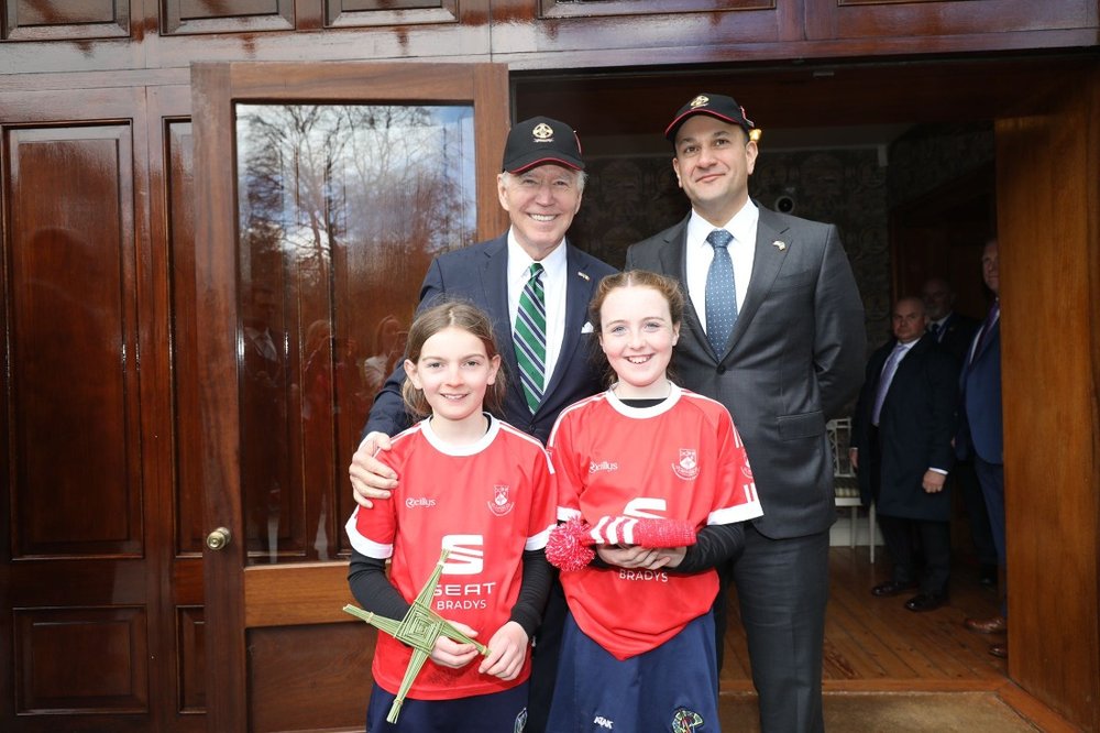 Two men wearing baseball hats standing behind two girls in their camogie uniforms holding a Brigid's cross