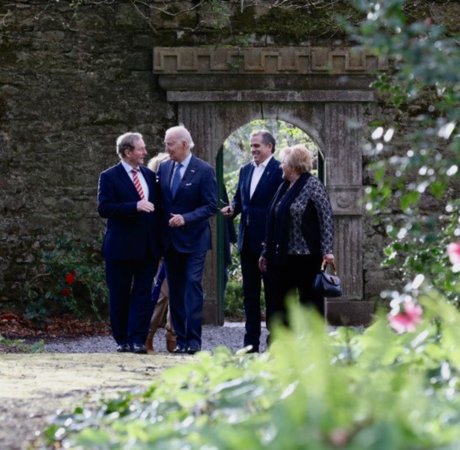 three men and one woman talking in a rose garden in front of an old wall
