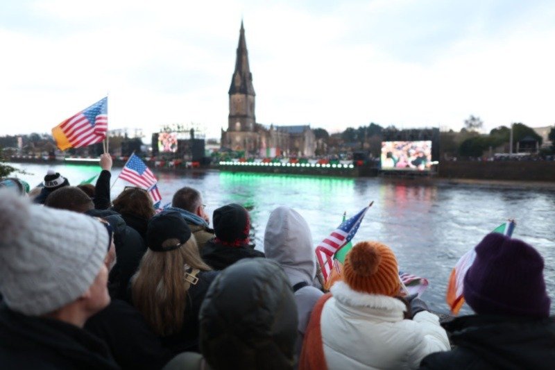 Church with spire across a dyed green river with people standing on the bank waving flags and cheering
