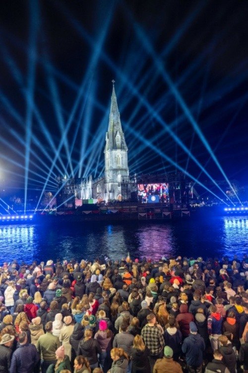 cathedral lit up with beams of light into the night sky above it with crowds facing it from across the river
