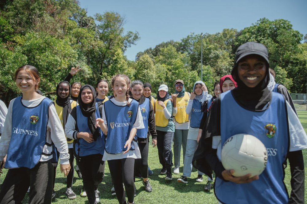 Teenage girls from multiple backgrounds walking across the pitch toward the camera. They are smiling in sports bibs and one is carrying a Gaelic football