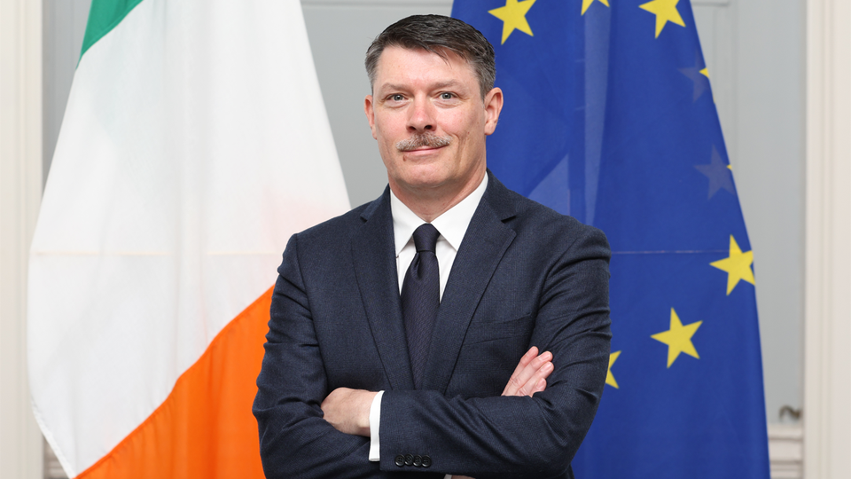 Ambassador Martin Gallagher standing in front of the Irish and EU Flags