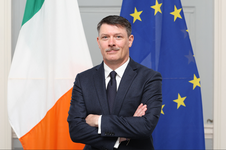 Ambassador Martin Gallagher standing in front of the Irish and EU Flags