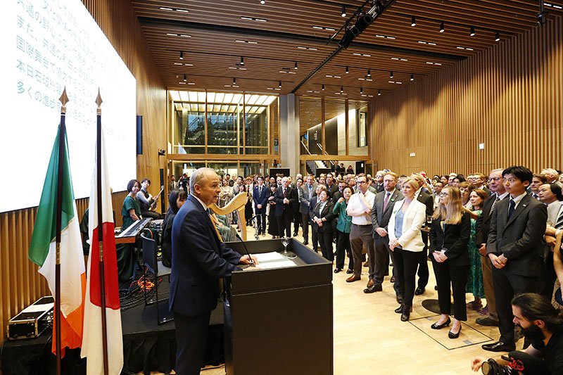 man addressing a crowd in Ireland House with flags behind him