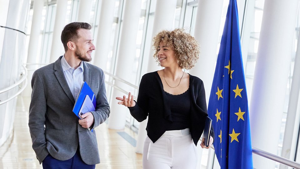 Two people walking and talking with an EU flag visible behind them