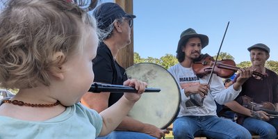A young girl playing the tin whistle along with adults playing the bodhran and the fiddle.