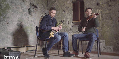 two men playing fiddle and uileann pipes in a stone room