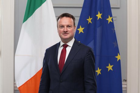 Portrait of Consul General Peter Neary standing in front of Irish and EU flags.