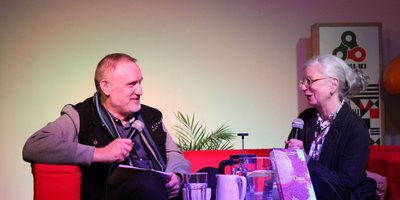 two people, a man and a woman, talking on a red sofa with microphones
