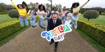 man in suit holding a sign with Africa Day surrounded by group of dancers with garden in the background