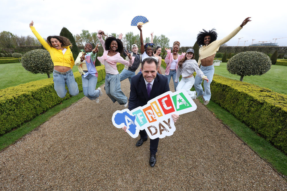 man in suit holding a sign with Africa Day surrounded by group of dancers with garden in the background
