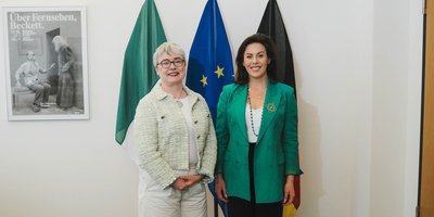 Ambassador Maeve Collins and Minister Jennifer Carroll MacNeill stand for a photo in front of large Irish, EU and German flags.