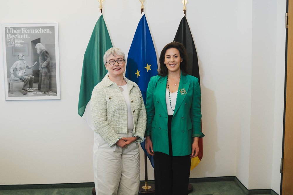 Ambassador Maeve Collins and Minister Jennifer Carroll MacNeill stand side by side for a photo in front of large Irish, EU and Germany flags.