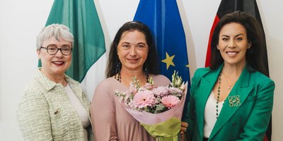 (L-R) Ambassador Maeve Collins, Professor Sabine Asmus and Minister Carroll MacNeill stand in front of large Ireland, EU and Germany flags. Professor Asmus holds a vase with a bouquet of pink flowers.