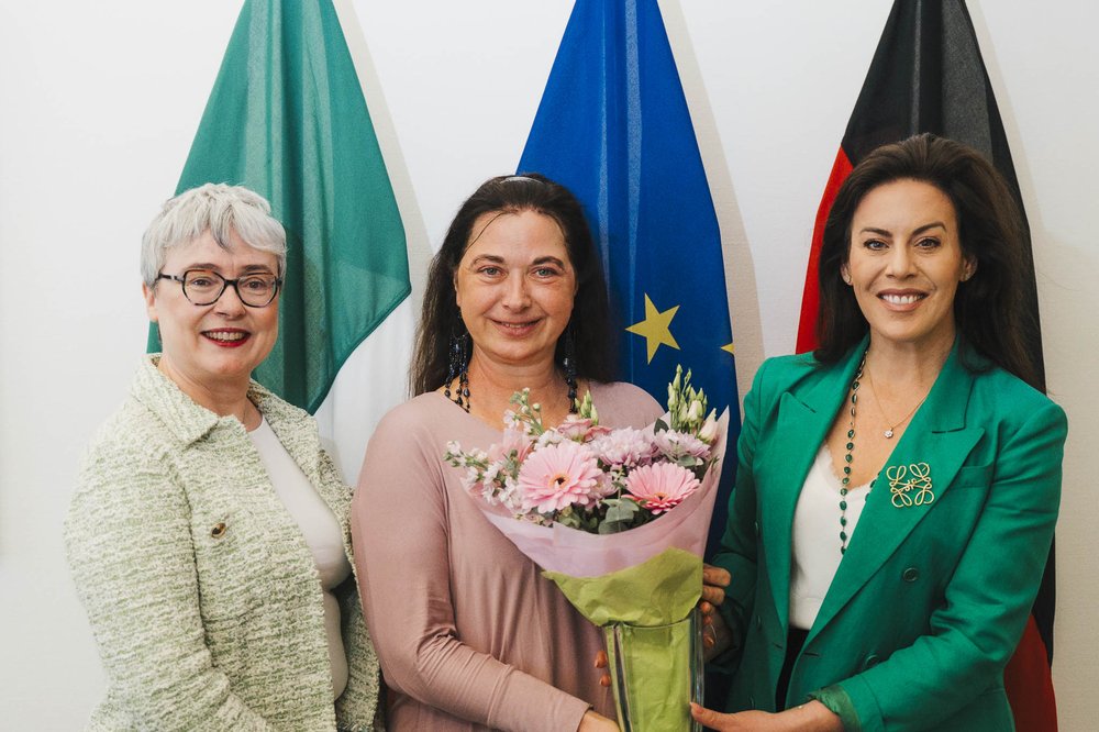 (L-R) Ambassador Maeve Collins, Professor Sabine Asmus and Minister Carroll MacNeill stand in front of large Ireland, EU and Germany flags. Professor Asmus holds a vase with a bouquet of pink flowers.
