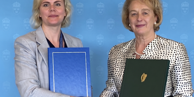 State Secretary of the Swedish Ministry for Finance Carolina Lindblom shaking hands with Ambassador Barbara Jones in front of a blue backdrop with Sweden's state emblem holding folders containing a newly signed treaty between the countries.