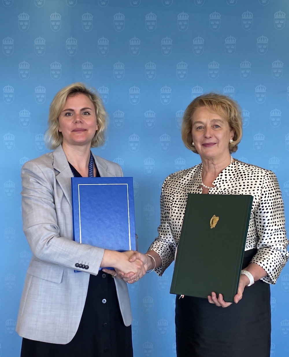 State Secretary of the Swedish Ministry of Finance Carolina Lindblom shaking hands with Ambassador Barbara Jones in front of a blue backdrop with Swedens state emblem holding folders containing a newly signed treaty between the countries.