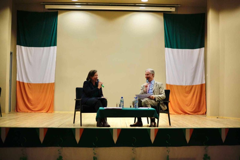 Two people on stage addressing an audience; Irish flags in the background