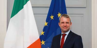 Shane Keegan in a smart suit in front of the Irish and EU flags