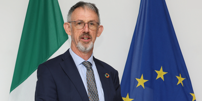 Ambassador Aidan Fitzpatrick standing in front of the Irish and EU flags.