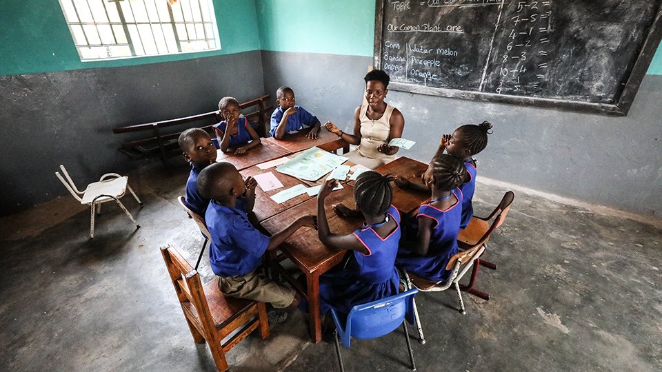 A teacher sits with a group of young pupils at a large table.
