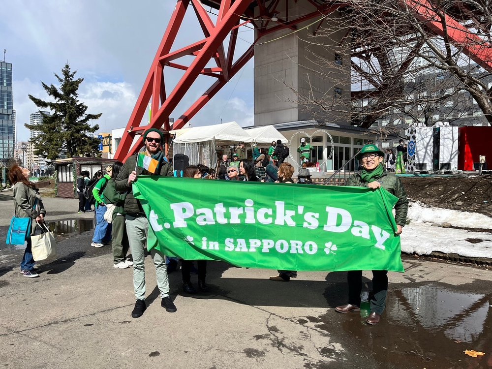 Two men holding a green banner reading "St Patrick's Day in Sapporo" both are smiling and wearing Irish themed accessories